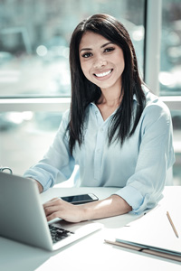 Professional woman smiling at camera while working on laptop in bright office setting