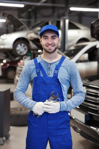 Male auto mechanic in blue overalls smiling and holding wrenches while standing among vehicles in a large garage