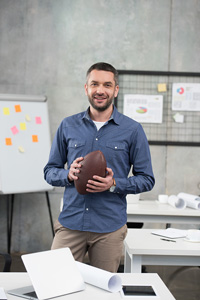 Smiling male sports agent in khakis and a button-down shirt holding a football and standing in a room with tables and charts