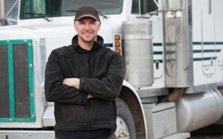 Truck driver in a black hoodie and ball cap stands in front of a white and green semi truck
