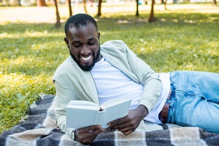Smiling young man reading a book outside in the shade while lying on a checkered blanket over grass