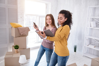 Two smiling young female college students looking at mobile devices in a bright room with moving boxes on the floor
