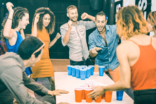 Several young men and women standing and celebrating around a white table during a game of beer pong with blue and red cups