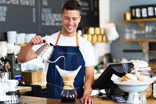 Smiling male barista at the counter of a coffee shop pouring water from a silver kettle into a filter over a coffee pot