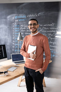 Smiling man wearing glasses, holding a digital tablet, and standing next to a desk with a computer and open laptop