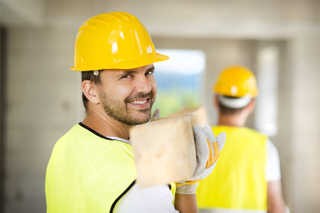 Smiling construction worker in yellow hard hat and high-visibility vest carrying lumber with colleague in background