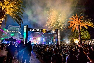 Crowd enjoying outdoor concert with fireworks display at Full Sail Studios event at night with palm trees.