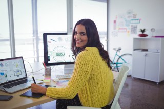 Young woman in yellow sweater doing design work on several computers at a desk