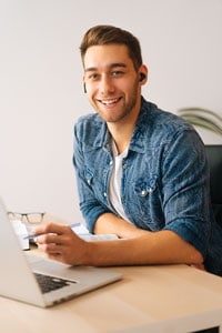 Young man sitting at a desk with a laptop, wearing wireless earbuds and smiling at the camera.