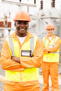 Two construction workers in orange safety vests and hard hats smiling at an industrial site