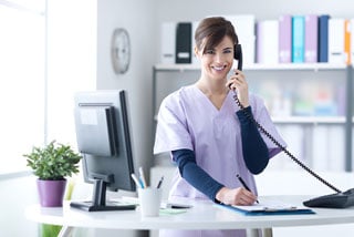 Smiling medical receptionist talking on phone while working at clinic desk with computer and office supplies