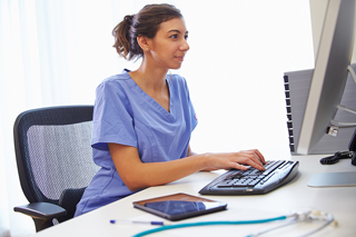 Woman in medical scrubs using computer at workstation with stethoscope and smartphone on desk