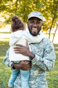 Smiling male service member in a camouflage military uniform standing outside and holding a young girl who is hugging him