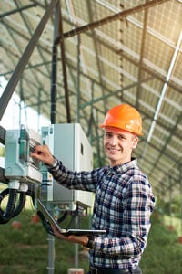 Male electrician in a plaid shirt and orange hard hat inspecting a piece of equipment beneath an array of solar panels