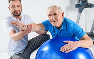 Older man leaning against a stability ball and extending one arm while another man kneels beside him and guides the arm