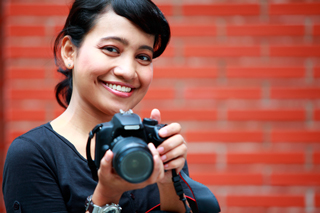 Smiling photographer holding a DSLR camera in front of a brick wall