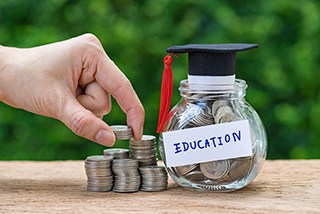 Hand stacking coins with a labeled glass jar for education savings topped with a graduation cap against a blurred green background
