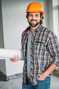 Smiling man in a plaid shirt and orange hard hat holding blueprints and standing inside a building under construction