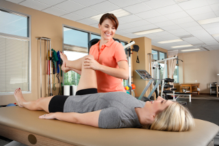 Woman performing a leg stretch exercise on another woman lying on a physiotherapy table in a well-lit rehabilitation gym.