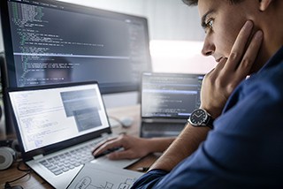 Side view of a focused male programmer working on multiple computer screens with code and development software
