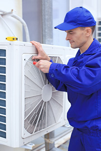 Man in a blue hat and technician uniform using a screwdriver to work on air conditioning equipment outside a building