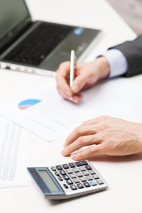 Closeup of hands writing on a document with pen, calculator and laptop on desk, business analysis concept.