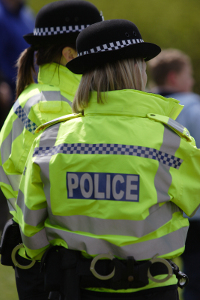 Two police officers wearing high-visibility jackets with "POLICE" written on them, viewed from the back at an outdoor event.