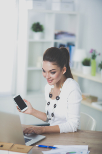 Administrative assistant in white blouse smiling at smartphone at her desk with laptop and decorative items in a bright office.