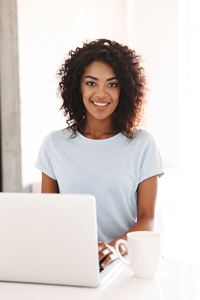 Young woman with curly hair smiling at camera while working on laptop at a bright home office
