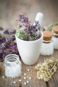 Mortar and pestle with fresh purple flowers, homeopathic pills, and jars of herbal medicine on wooden table