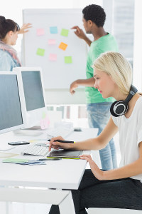 Young blonde woman with headphones using a computer in a busy office, with colleagues discussing at a whiteboard in the background.