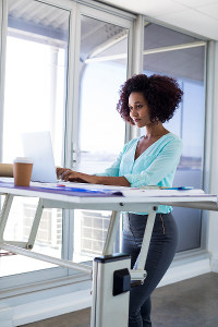 Female architect standing at adjustable desk working on laptop in bright office with city view