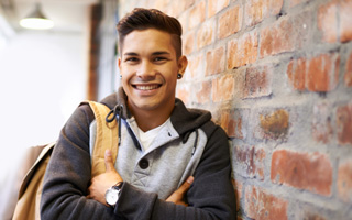 Smiling young man leaning against a brick wall with a backpack and wearing a casual hoodie and watch.