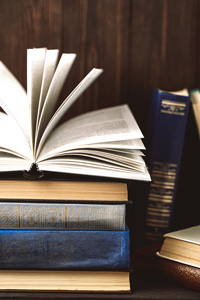 Open book on top of a stack of various hardcover books on a wooden table with a dark background