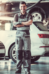 Male mechanic in gray uniform standing in automobile workshop with cars in the background.