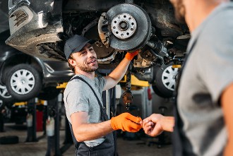 Mechanic in a cap and gloves works on a car's brake system while talking to a coworker in an automotive workshop.
