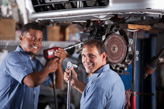 Two mechanics working on a car in a garage, one using a pneumatic wrench and the other using a socket wrench.