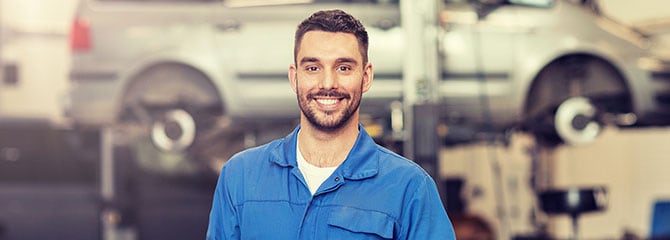 Smiling young man in blue coveralls holding a digital tablet and standing in front of a car hoisted on an automotive lift