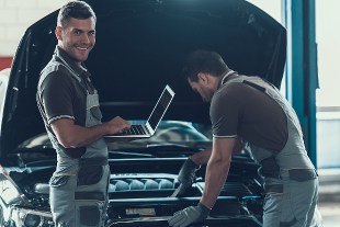Two mechanics working on a car engine in a garage, one holding a laptop and smiling at the camera.