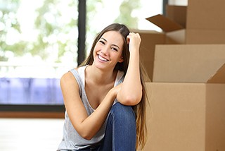 Young woman smiling and sitting on the floor near cardboard boxes in a sunlit room, possibly during a moving day.