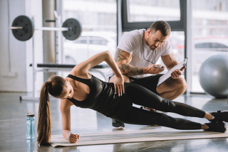 Woman performing a side plank exercise in a gym with assistance from a male trainer holding a clipboard