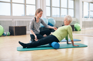 Elderly woman performing a stretch on a foam roller with assistance from a personal trainer in a gym with exercise balls in background.