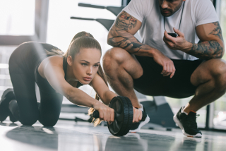 Woman performing a plank exercise with a dumbbell in a gym while a male personal trainer with tattoos uses a smartphone to time her.