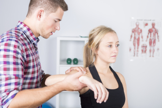 Physical therapist examining patient's arm in clinic with anatomy poster in background.