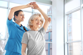 Elderly woman doing physical therapy exercises with a female therapist in a well-lit room with large windows.