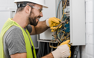 Smiling electrician wearing a blue hard hat and plaid shirt working on an electrical panel with a multimeter in hand.