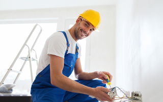 Smiling electrician in blue overalls and yellow hard hat wiring a socket in a bright room