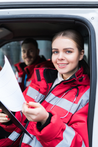Young female paramedic in red jacket holding paperwork and smiling in ambulance with male colleague in background