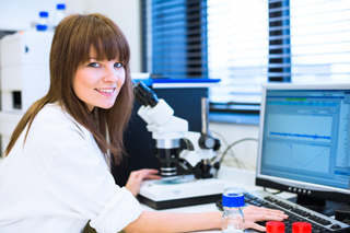 Female scientist with brown hair using a microscope and computer in a laboratory setting.