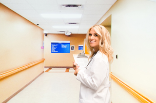 Blonde health care professional smiling and holding a clipboard in a hospital corridor
